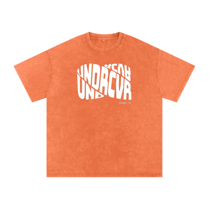 An orange mineral-wash t-shirt with a white graphic print on the front reading 'UNDRCVR' and 'BEYOND UNDRCVR' in a stylized overlapping layout.