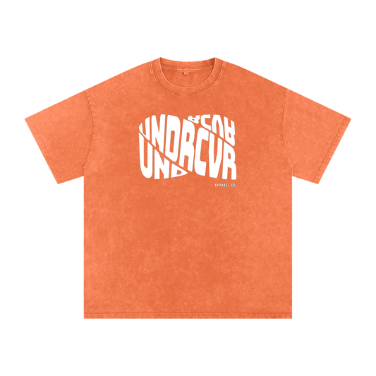 An orange mineral-wash t-shirt with a white graphic print on the front reading 'UNDRCVR' and 'BEYOND UNDRCVR' in a stylized overlapping layout.
