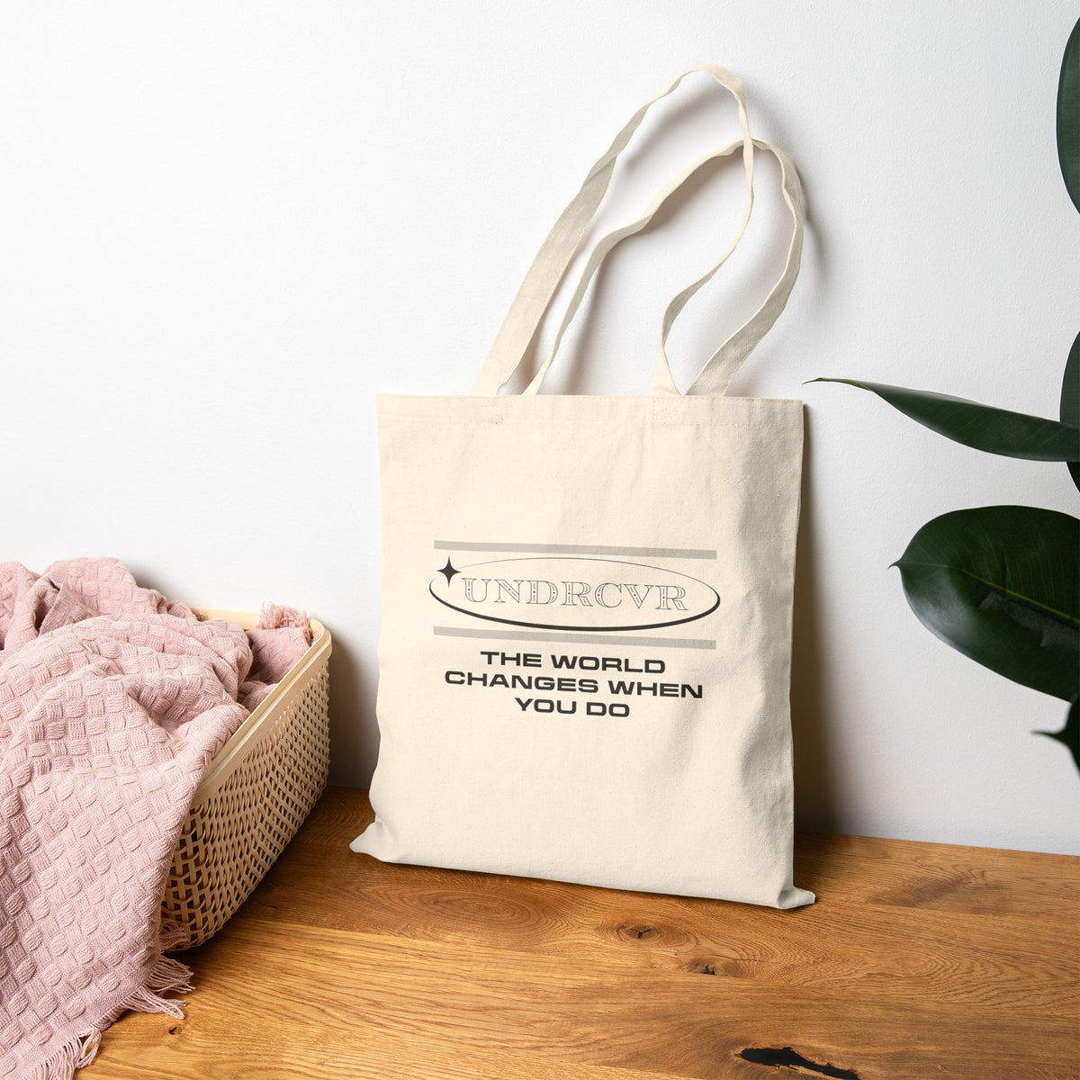 Beige tote bag with 'The World Changes When You Do' text on a wooden surface with a white wall and plant in the background.