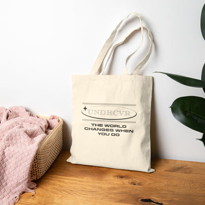 Beige tote bag with 'The World Changes When You Do' text on a wooden surface with a white wall and plant in the background.