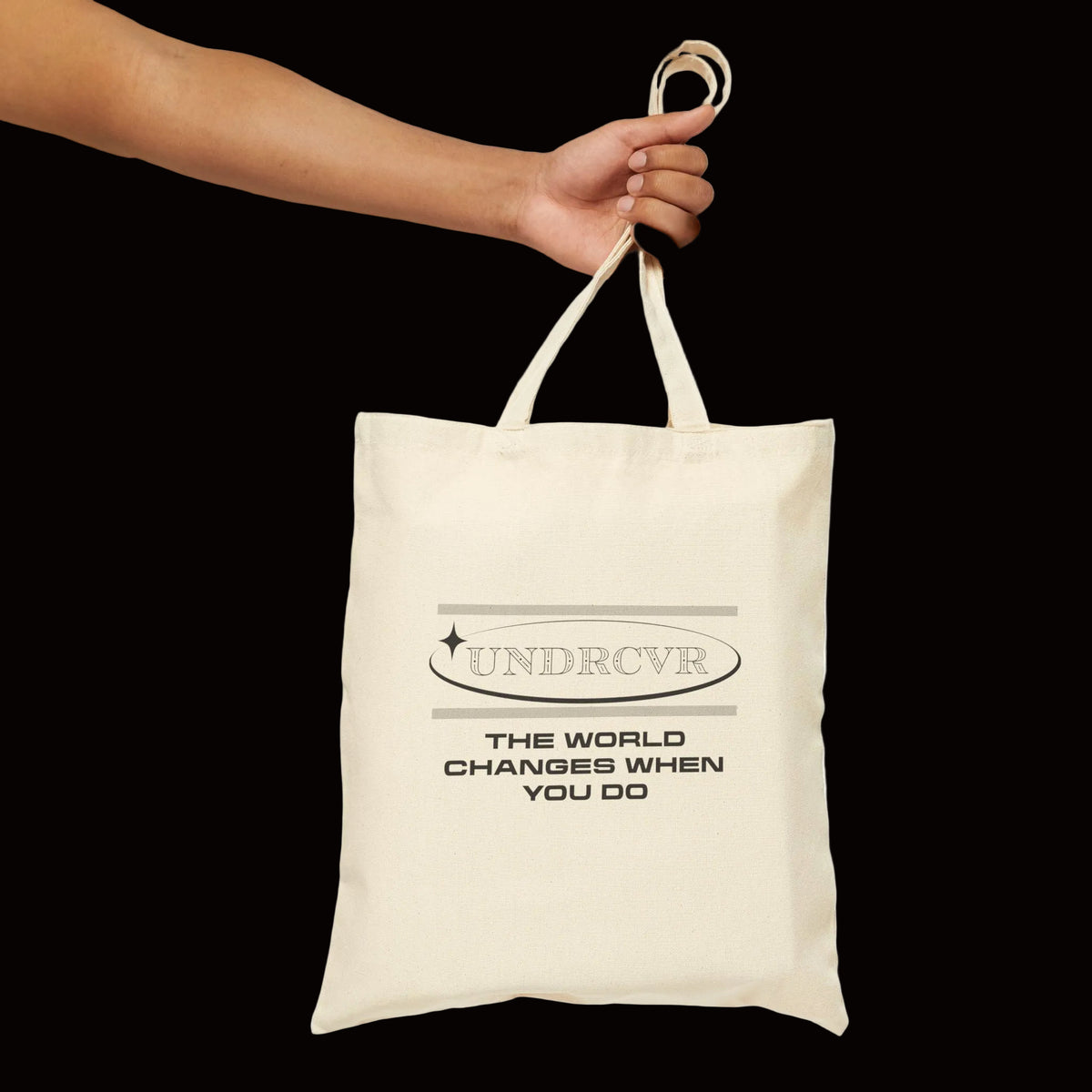An off-white canvas tote bag with black printed text reading 'UNDRCVR THE WORLD CHANGES WHEN YOU DO' being held by a person's hand against a plain white background.