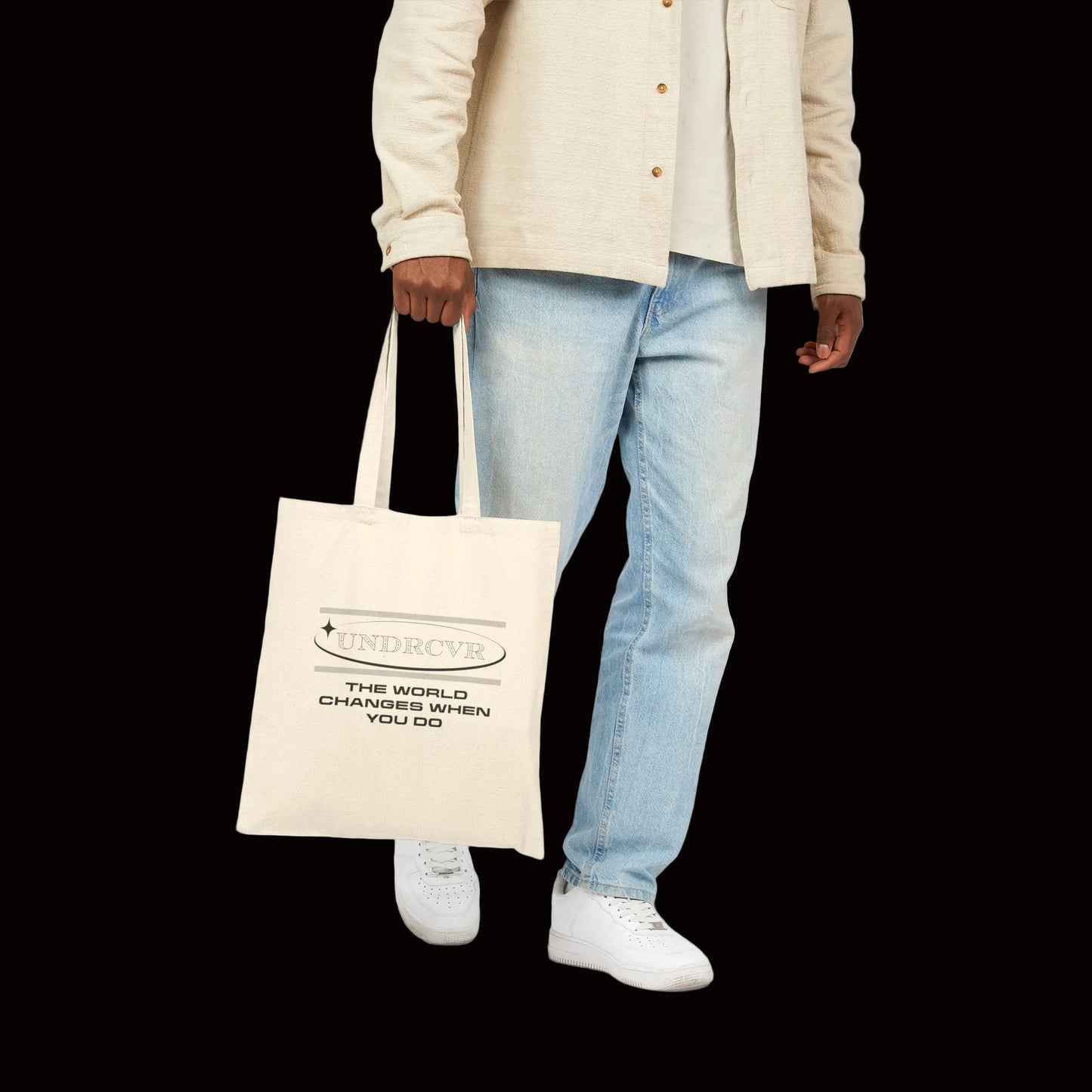 A person wearing a beige button-up shirt, light blue jeans, and white sneakers holds a cream canvas tote bag with black text that reads 'UNDCVR' and 'THE WORLD CHANGES WHEN YOU DO'.
